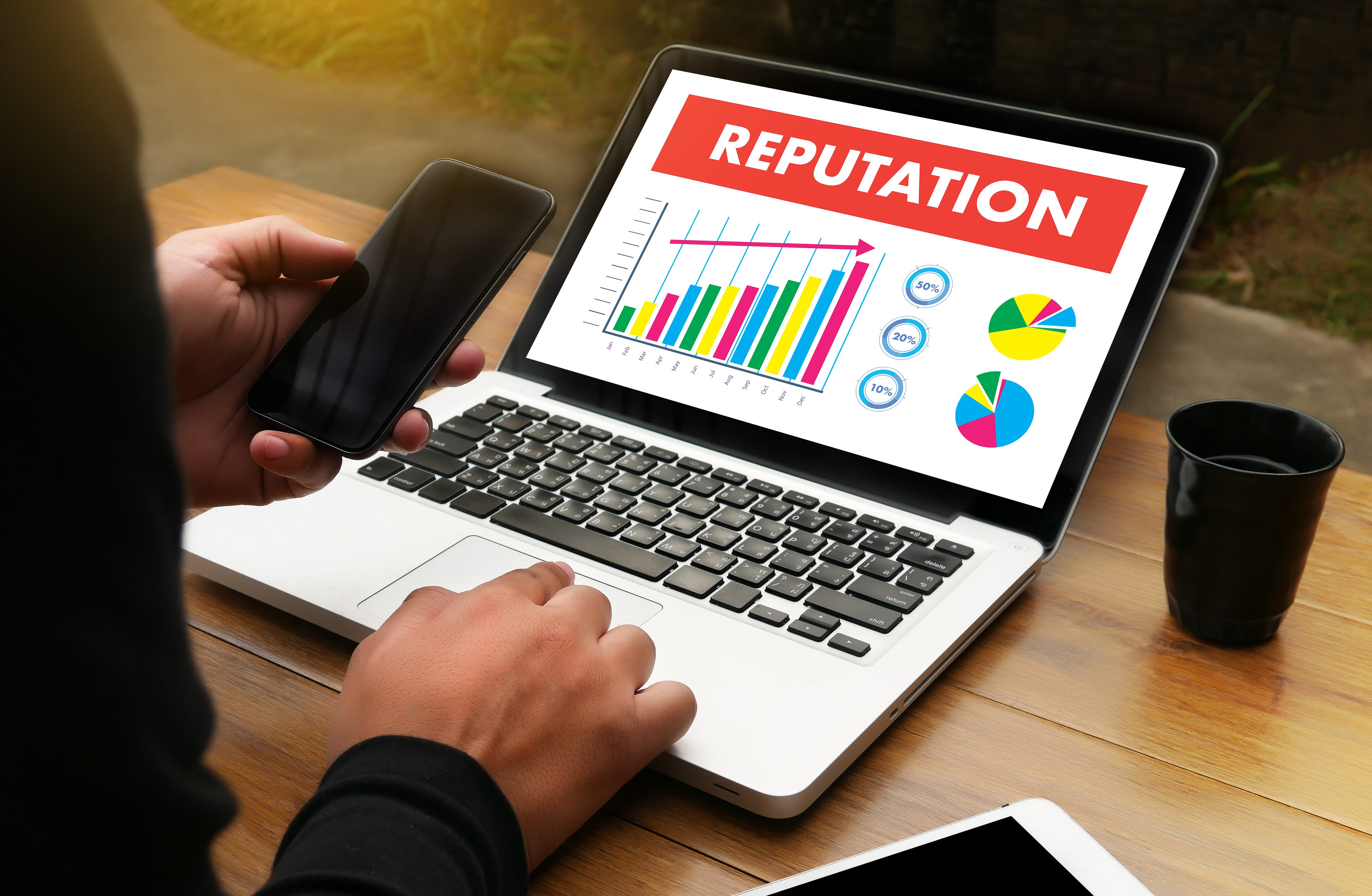 Image of an individual in a business suit, working on their laptop, at a desk. On the individuals laptop, there is an image. The image on the laptop, is a bar graph. The bar graph is showing an increase in online reputation. 