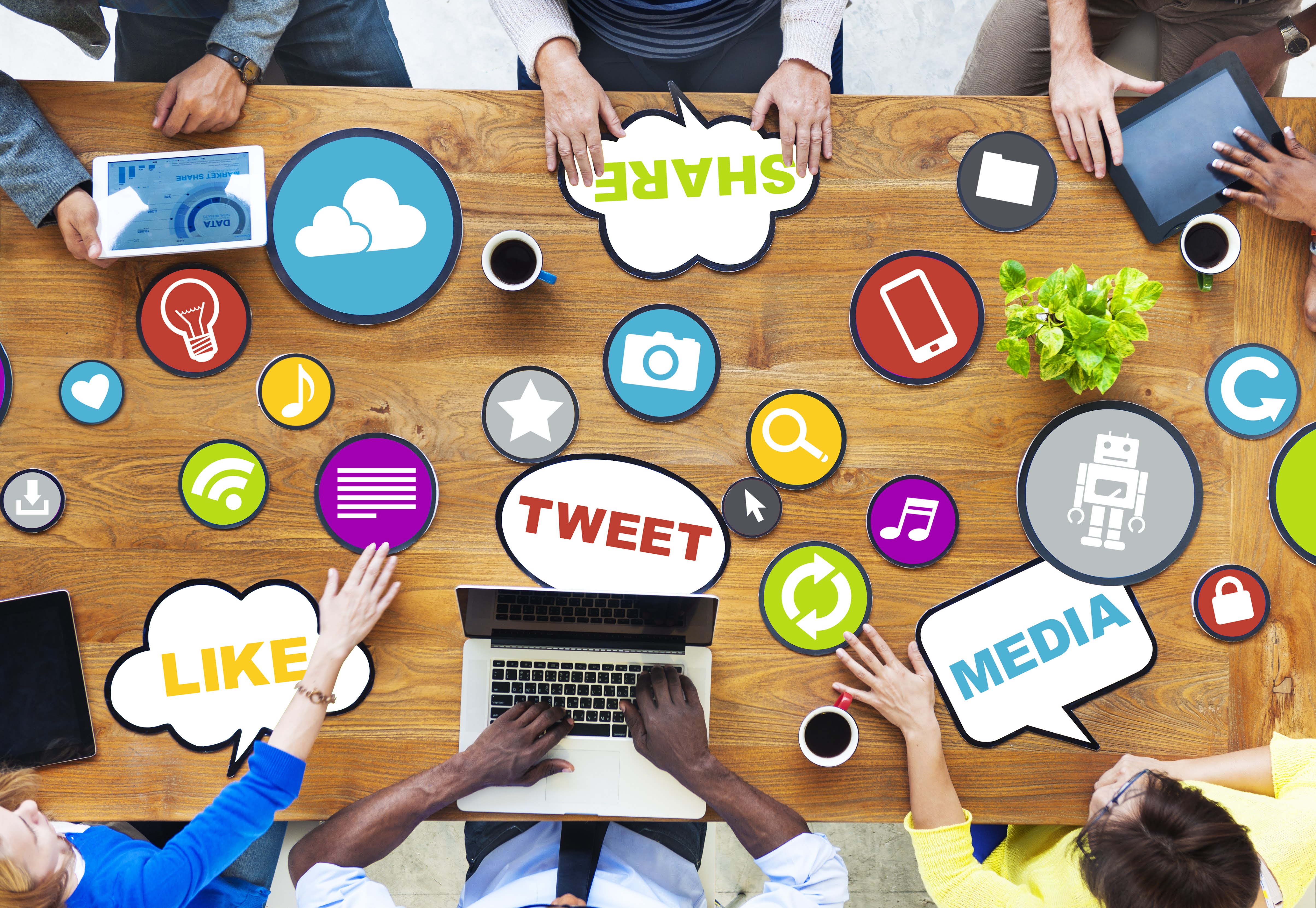 Image of six people at a table. On the table there is multiple electronic devices. There is multiple  comment bubbles on the table; which demonstrate a brain storming session. The individuals at the table are brainstorming an optimal social media marketing campaign. 